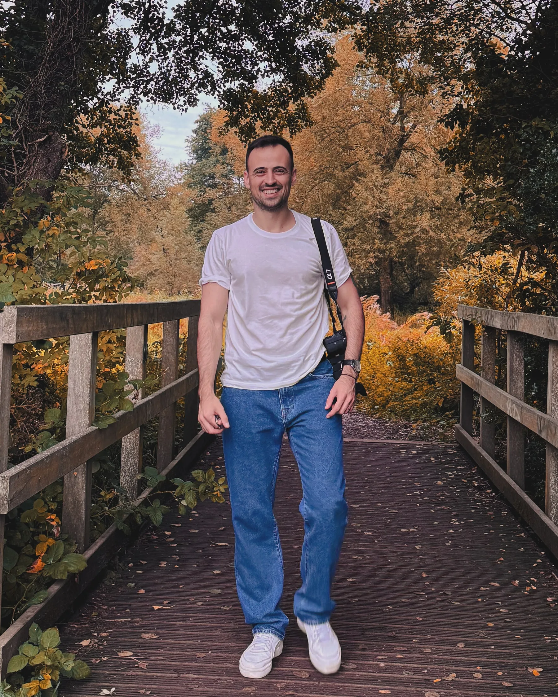 Can Ozturk, wedding and portrait photographer, standing on a wooden footbridge through autumn woodland with a camera over his shoulder