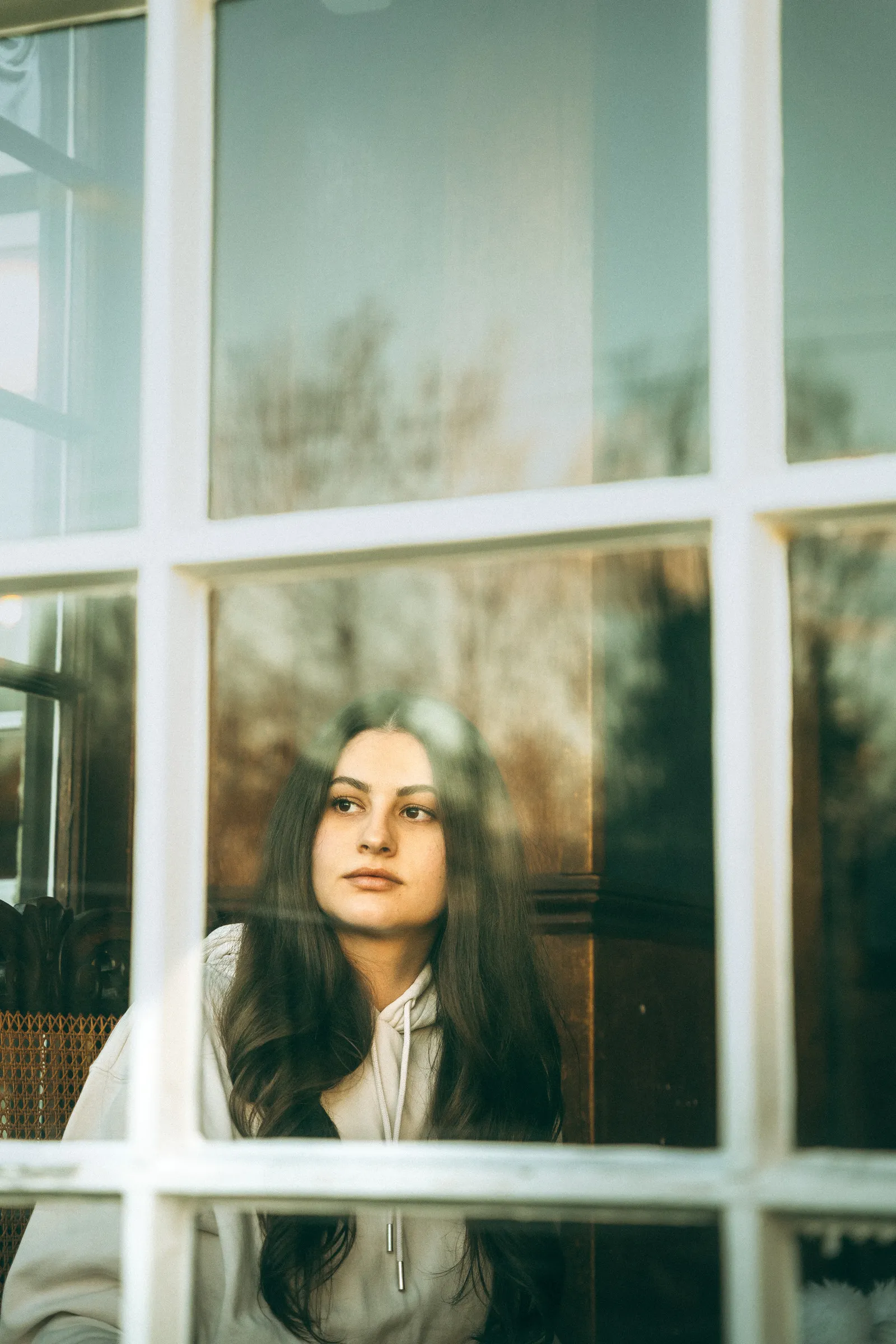 A woman looking out through a white sash window, autumn trees reflected in the glass