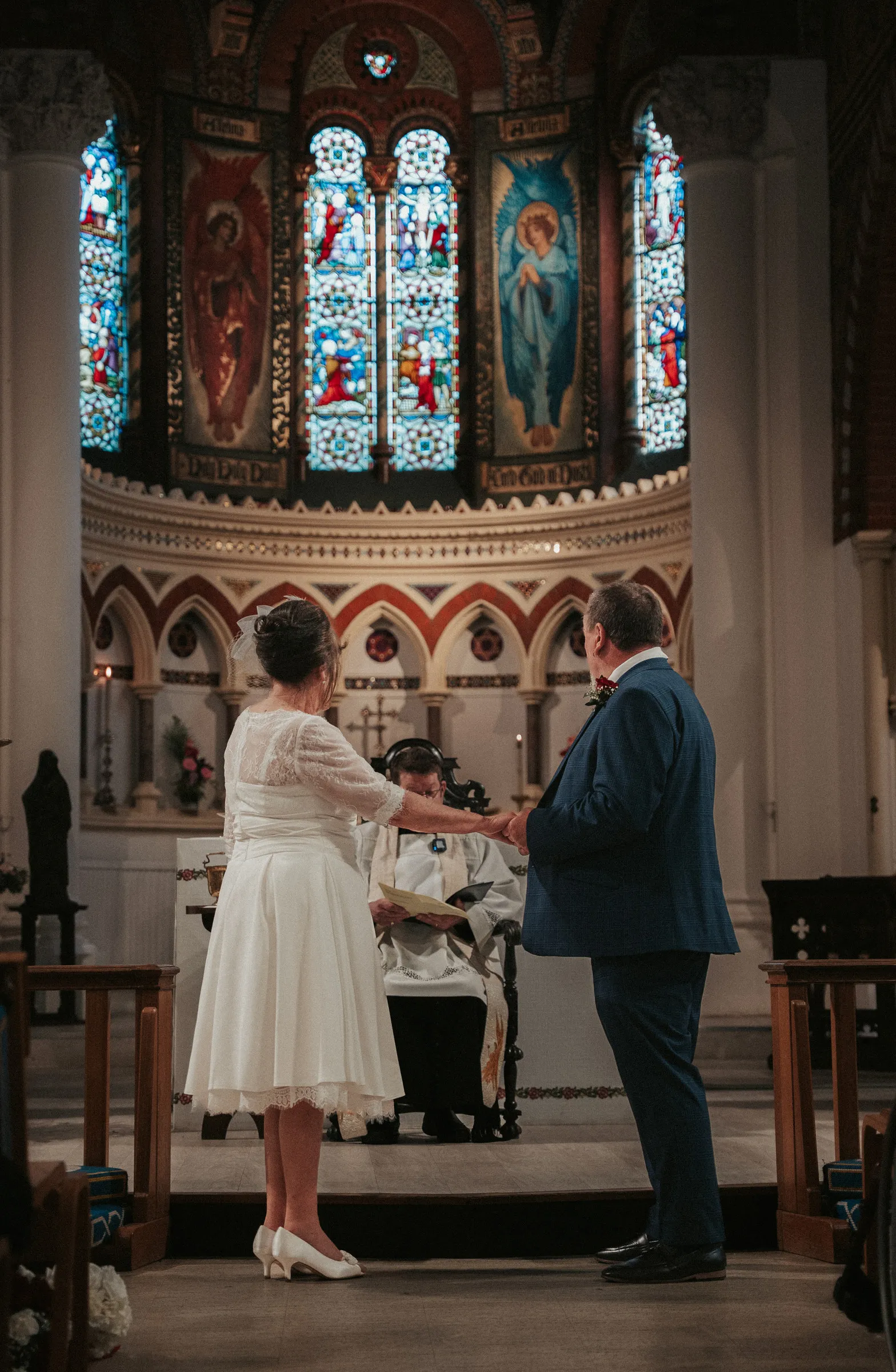 The couple standing side by side at the altar beneath stained glass